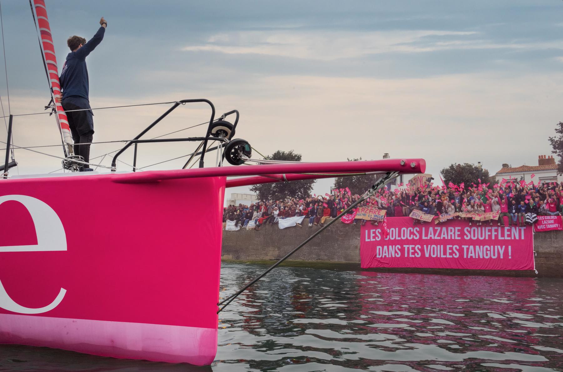 Skipper saluant la foule sur l’étrave rose du voilier Lazare, banderole de supporters en arrière-plan.
