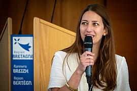 Femme brune parlant au micro devant un pupitre Région Bretagne, mise au point sur le visage, lumière de salle.