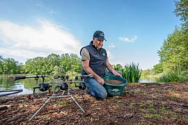 Technicien avec seau de granulés Le Gouessant et cannes à pêche sur la rive boisée d’un lac en Bretagne.