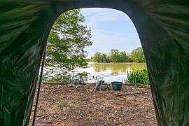 Ouverture d’une tente de bivouac sur un lac avec cannes à pêche sur rod pod, seau et végétation aquatique.