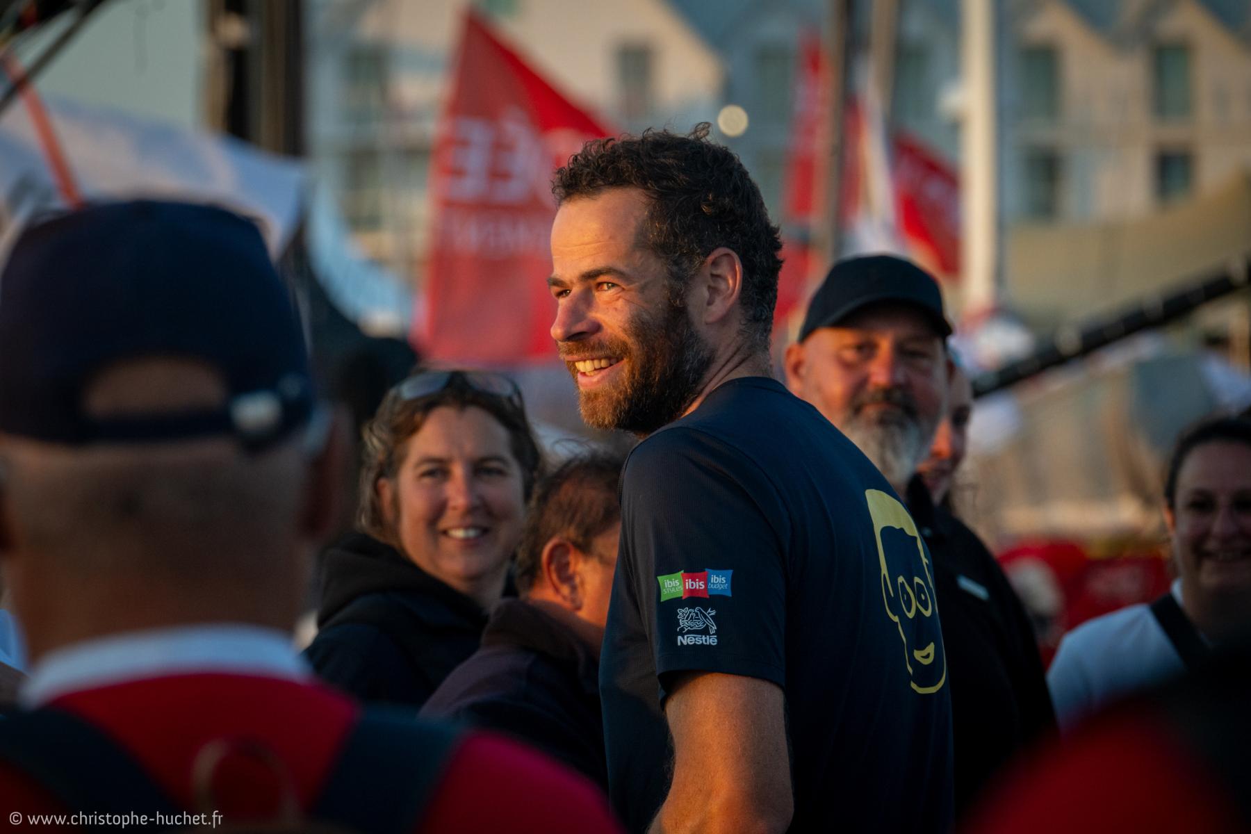 Portrait de Nico d’Estais barbu souriant en t-shirt bleu sous une lumière dorée lors de l’arrivée de la Vendée Arctique 2026