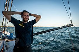 Marin barbu souriant sur son voilier en mer, portant un t-shirt Café Joyeux sous un ciel bleu et lumière du soir.