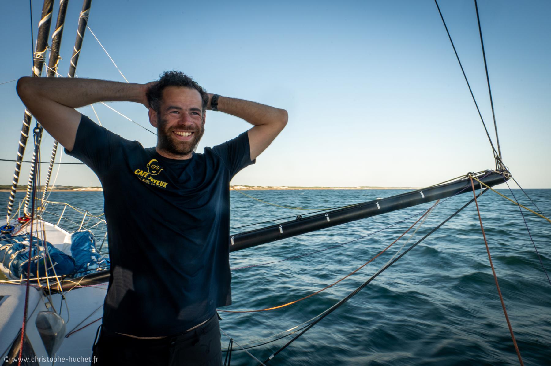 Marin barbu souriant sur son voilier en mer, portant un t-shirt Café Joyeux sous un ciel bleu et lumière du soir.