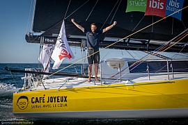 Marin bras levés sur le pont du voilier jaune Café Joyeux, mer bleue et drapeaux de course