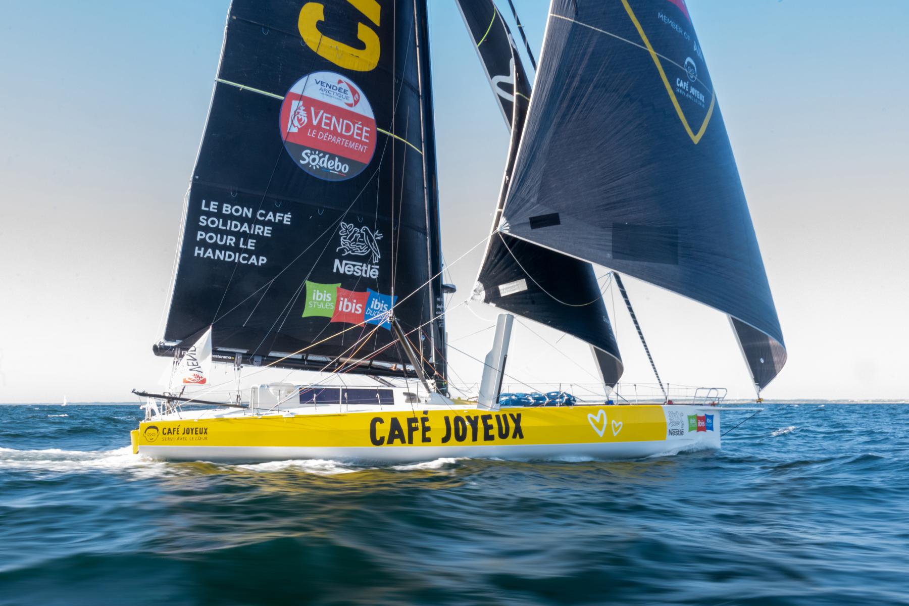 Imoca jaune Café Joyeux naviguant toutes voiles dehors sur une mer bleue par temps ensoleillé