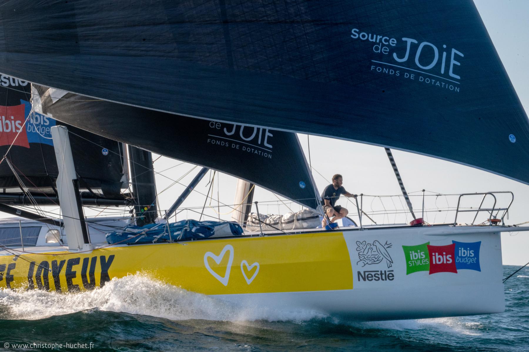 Voilier de course jaune Café Joyeux naviguant sur l’océan avec voiles noires déployées et un équipier sur le pont.