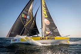 Voilier IMOCA Café Joyeux jaune et blanc fendant l’écume, voiles noires hissées sur une mer calme et ensoleillée.