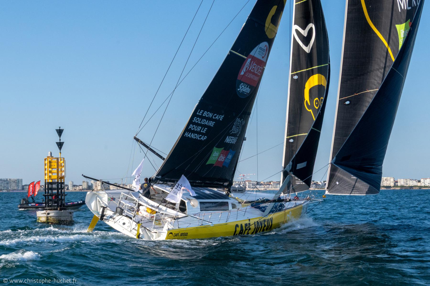 Imoca jaune et noir, Café Joyeux passant une bouée Cardinal Sud devant les Sables-d’Olonne, sous un ciel bleu ensoleillé de fin de journée