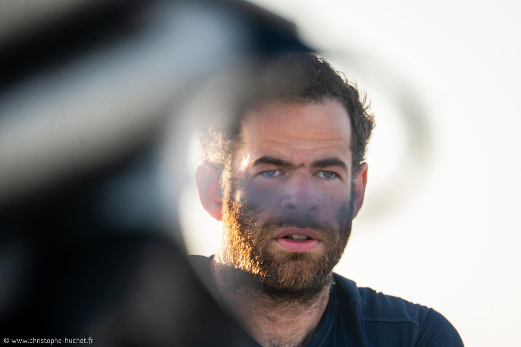 Portrait serré d’un skipper barbu au regard intense, visage marqué par l’effort, flou de premier plan, lumière vive.