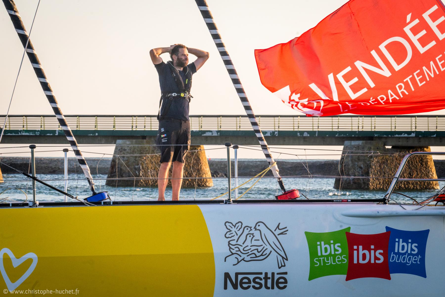 Skipper sur voilier jaune et blanc avec drapeau rouge au coucher du soleil devant la digue des Sables-d’Olonne