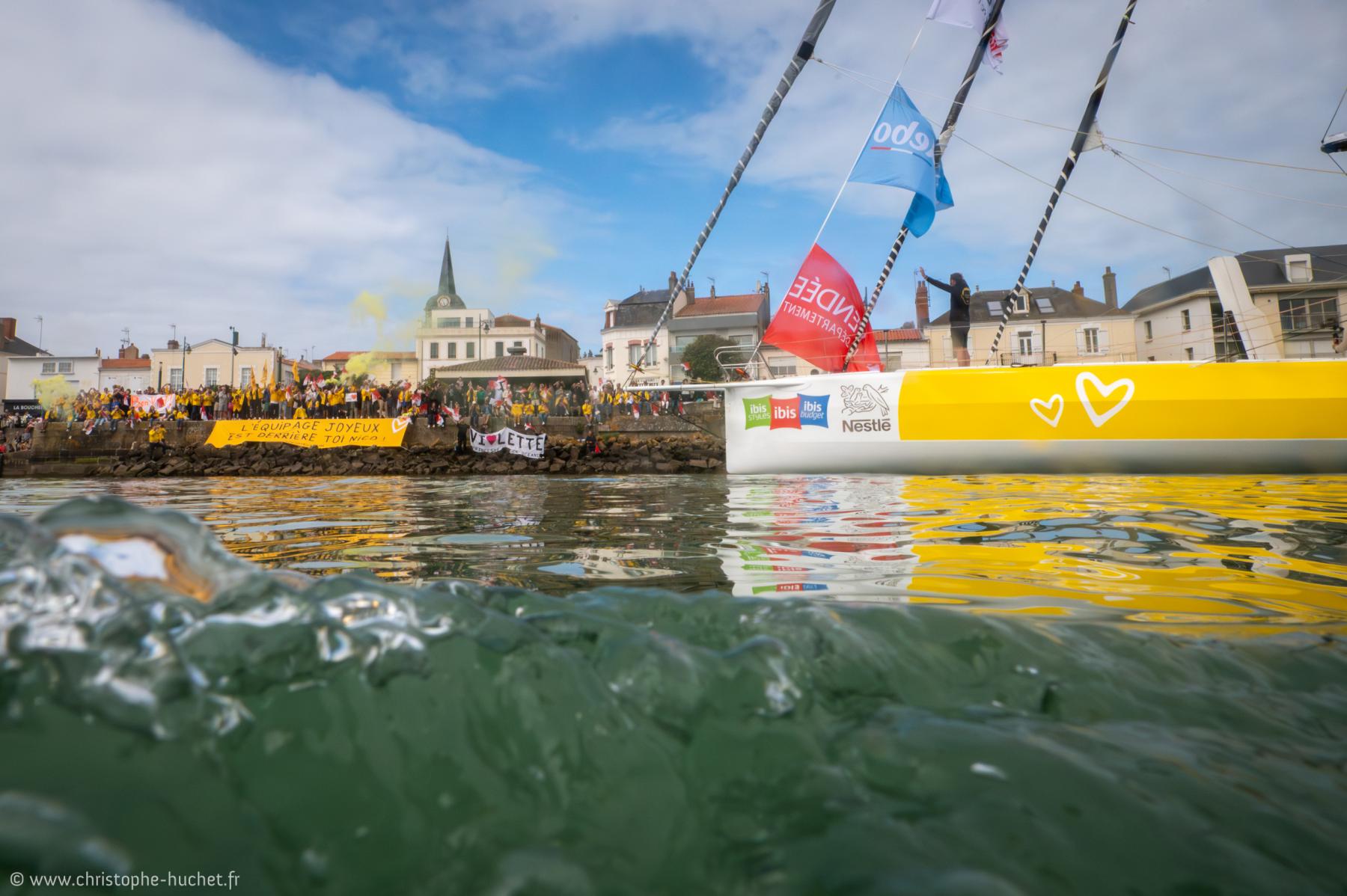 Coque jaune du voilier Café Joyeux vue au ras de l’eau. Foule massée sur le quai sous un ciel nuageux en Vendée.