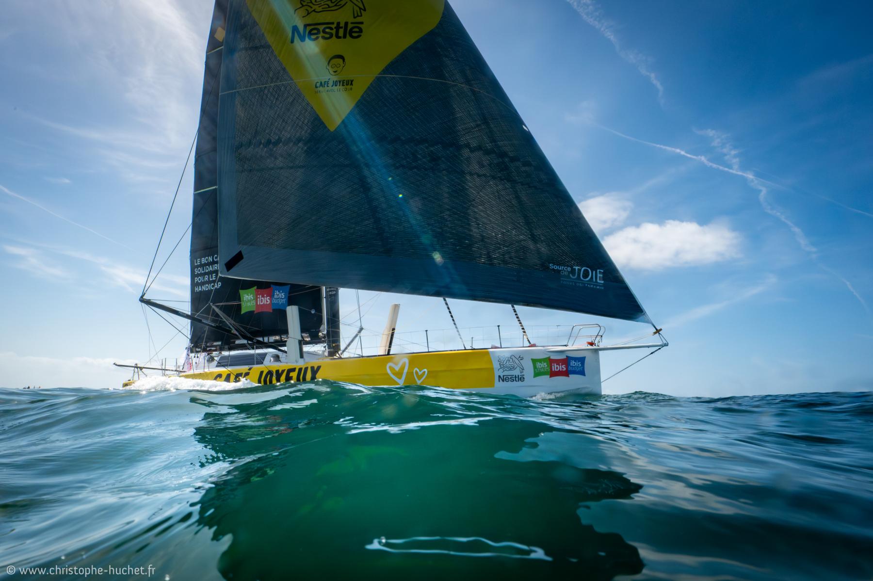 Voilier jaune Café Joyeux au ras de l’eau sur une mer calme et transparente, sous un ciel bleu avec trainées de nuages.