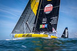 Voilier jaune Café Joyeux avec voiles noires sur l’océan, compétition nautique IMOCA sous un ciel bleu dégagé.