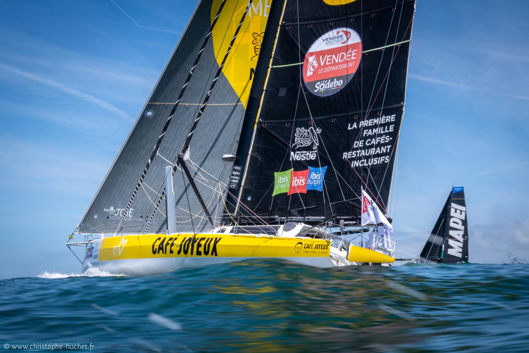 Voilier jaune Café Joyeux avec voiles noires sur l’océan, compétition nautique IMOCA sous un ciel bleu dégagé.