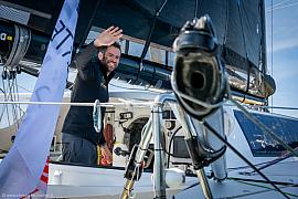 Skipper souriant saluant sur le pont du voilier Café Joyeux, gréement et bôme visibles sous un ciel bleu lumineux.