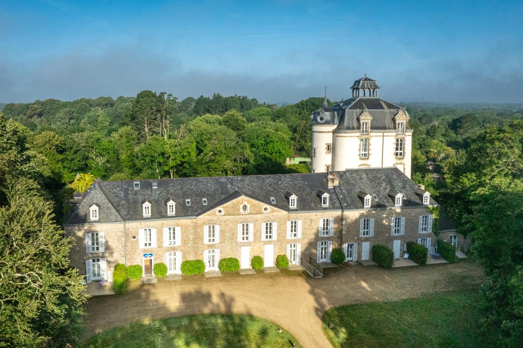 Vue aérienne d’un château en pierre claire, toit en ardoise et tour ronde, entouré d’une forêt dense sous un ciel bleu.