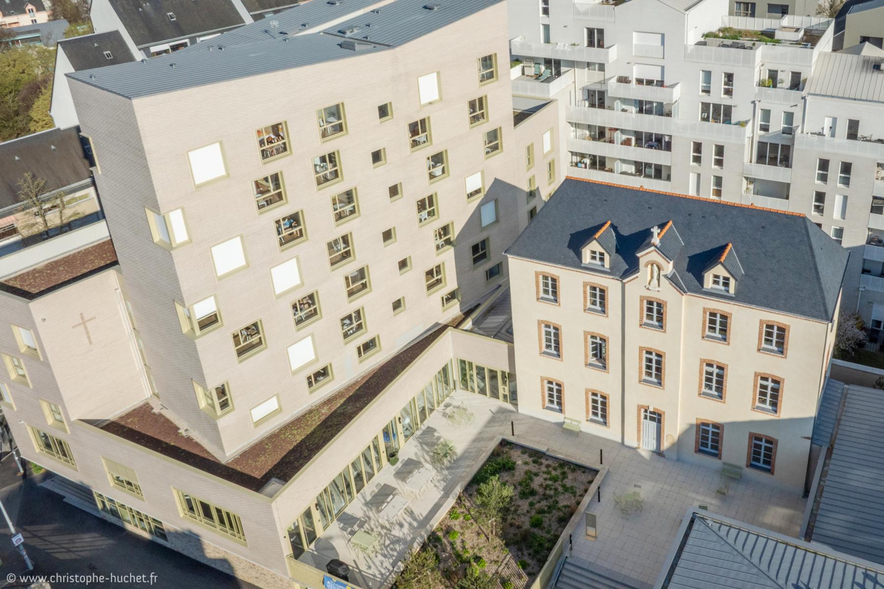 Vue de haut d’un bâtiment moderne beige aux fenêtres asymétriques jouxtant une bâtisse classique sous un soleil matinal.