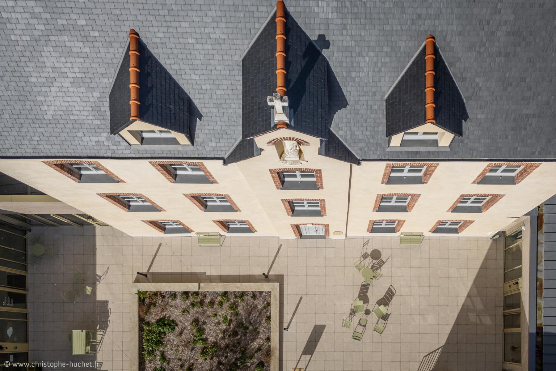 Vue verticale drone sur un toit en ardoise, facade beige et cour pavée ensoleillée avec mobilier vert.