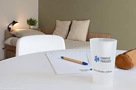 Bureau blanc avec gobelet logoté et bloc-notes au premier plan d’une chambre étudiante moderne et lumineuse à Rennes.