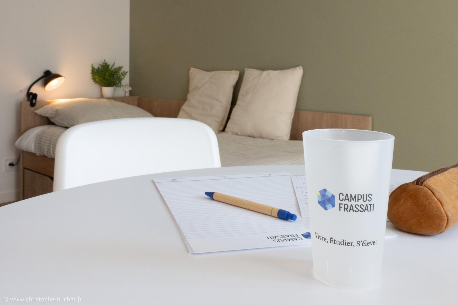 Bureau blanc avec gobelet logoté et bloc-notes au premier plan d’une chambre étudiante moderne et lumineuse à Rennes.
