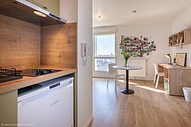 Kitchenette en bois au premier plan ouvrant sur un séjour lumineux avec bureau, table ronde et mur de cartes postales.
