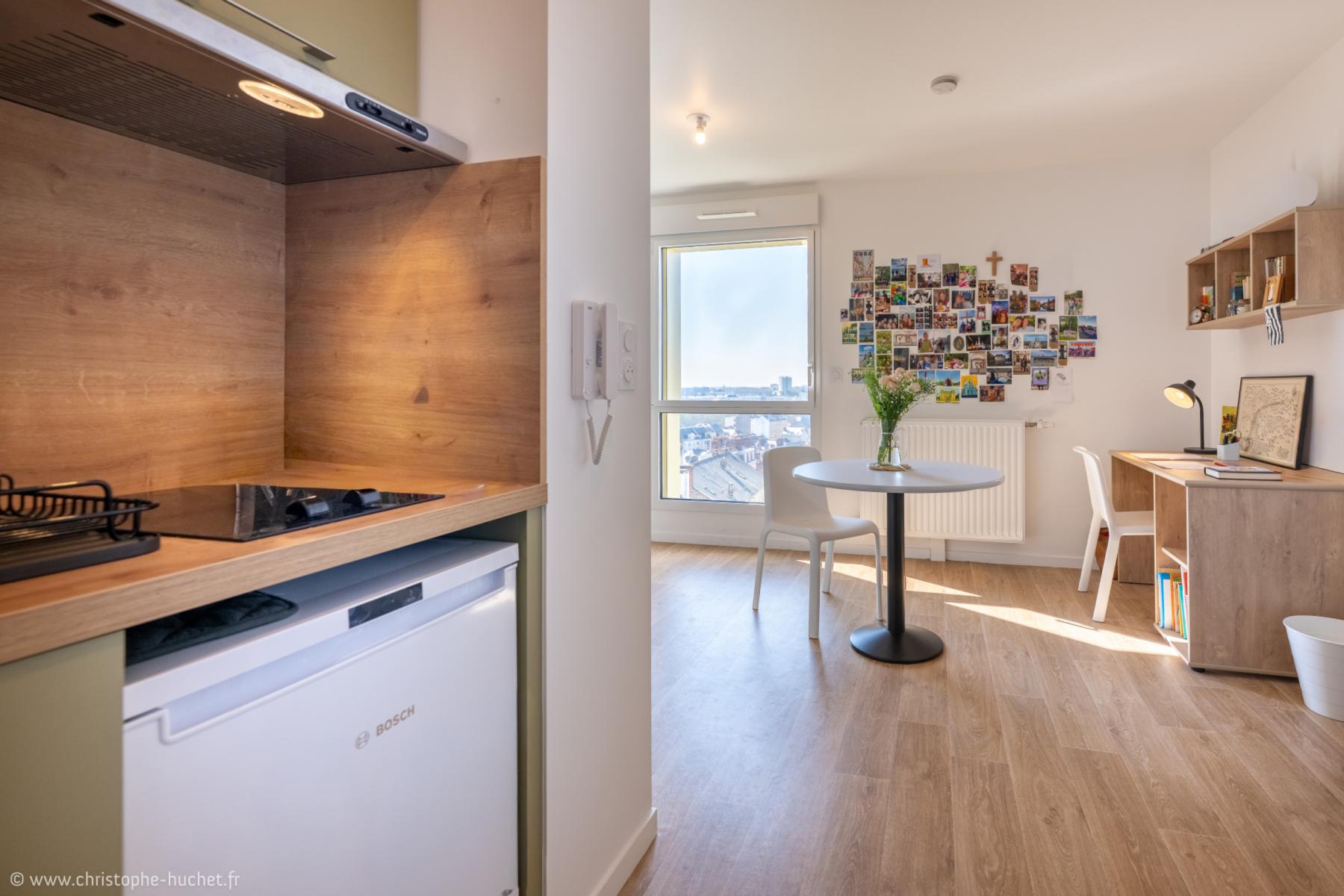 Kitchenette en bois au premier plan ouvrant sur un séjour lumineux avec bureau, table ronde et mur de cartes postales.