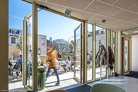 Vue intérieure vers une terrasse ensoleillée, portes vitrées ouvertes, étudiants attablés et silhouette en mouvement.
