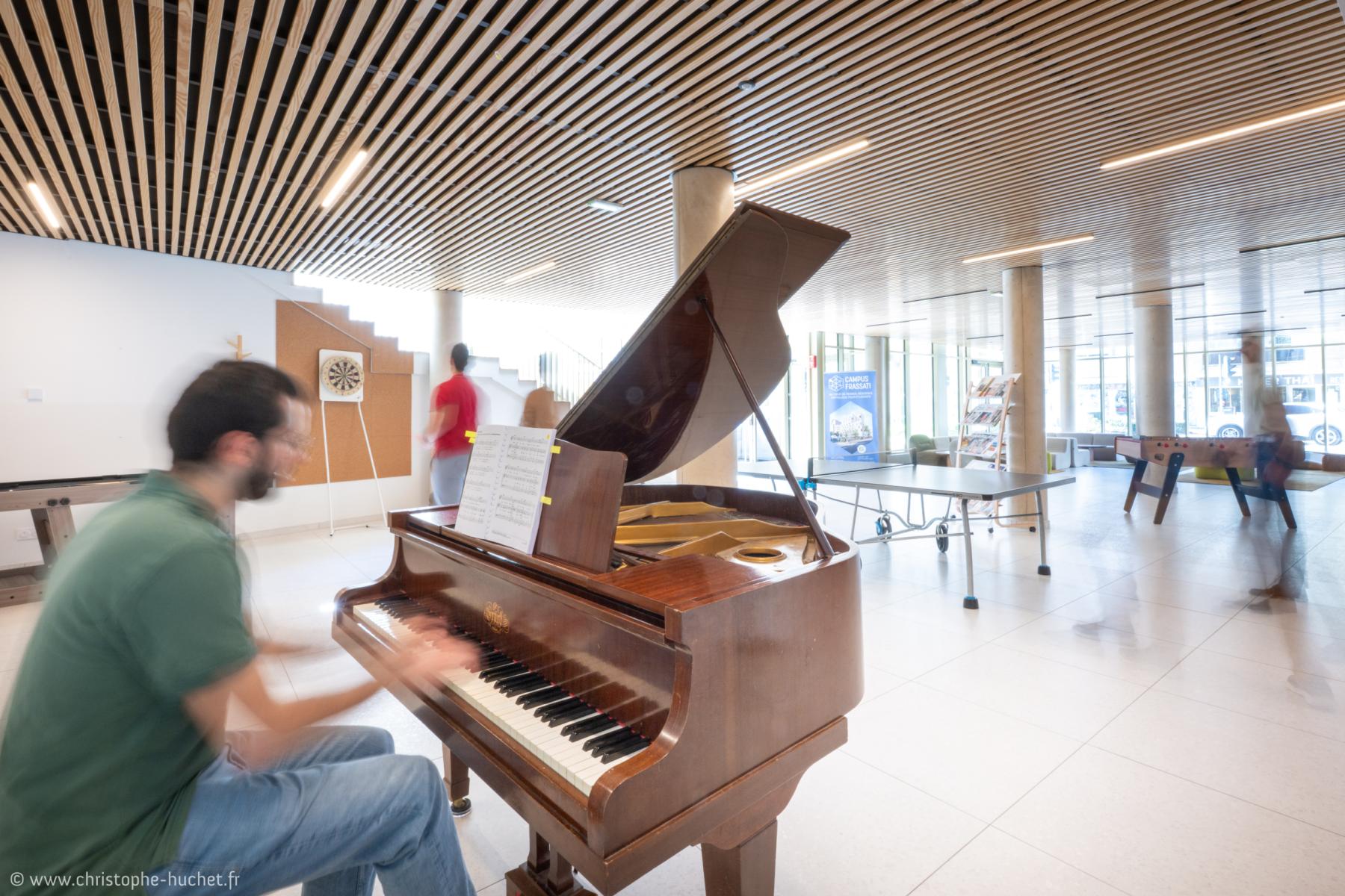 Homme jouant du piano à queue sous un plafond en lattes de bois, espace moderne lumineux avec tables de jeu.