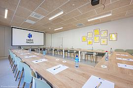 Salle de conférence en U avec tables en bois clair, chaises colorées, écran géant et décoration murale jaune.