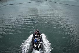 Bateau semi-rigide noir transportant un groupe en mer, vue de dessus avec sillage blanc sur l’eau sombre.