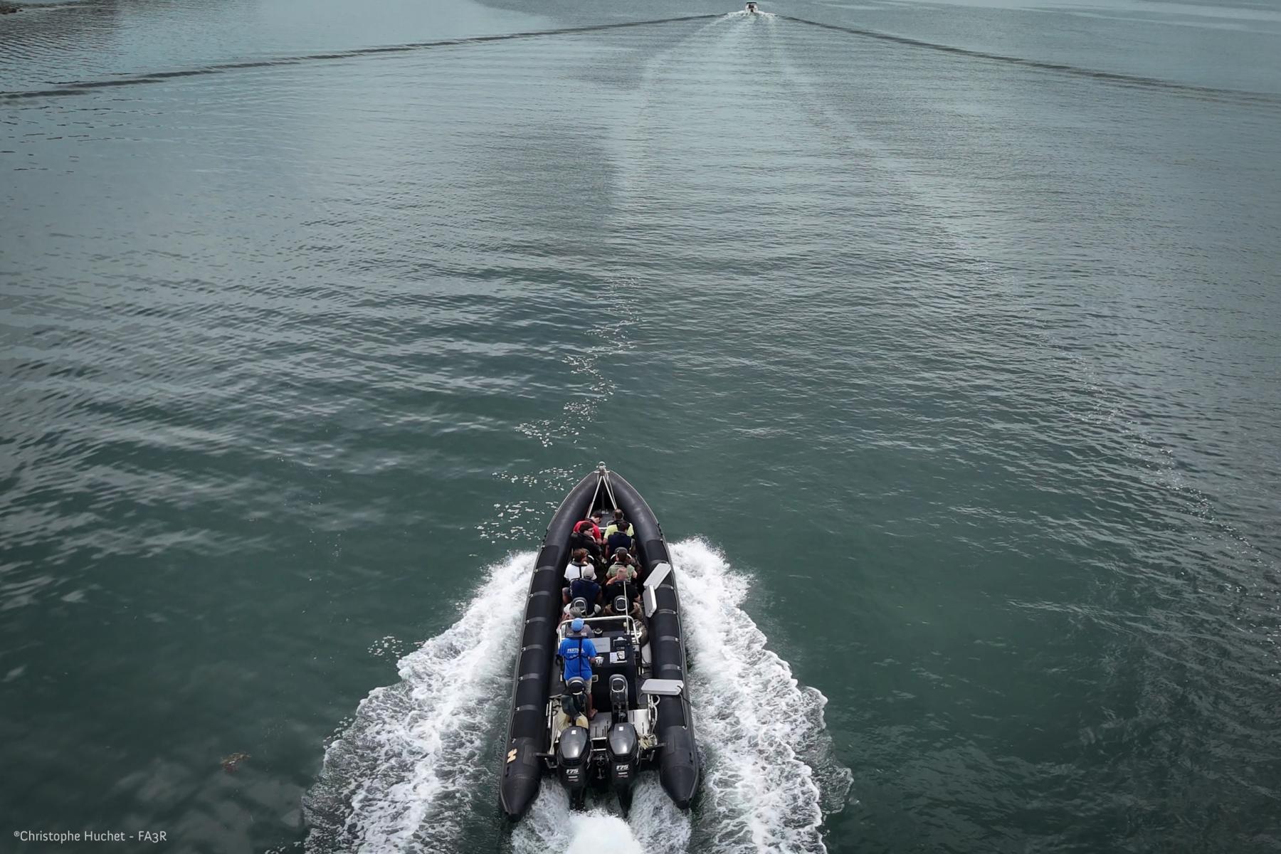Bateau semi-rigide noir transportant un groupe en mer, vue de dessus avec sillage blanc sur l’eau sombre.