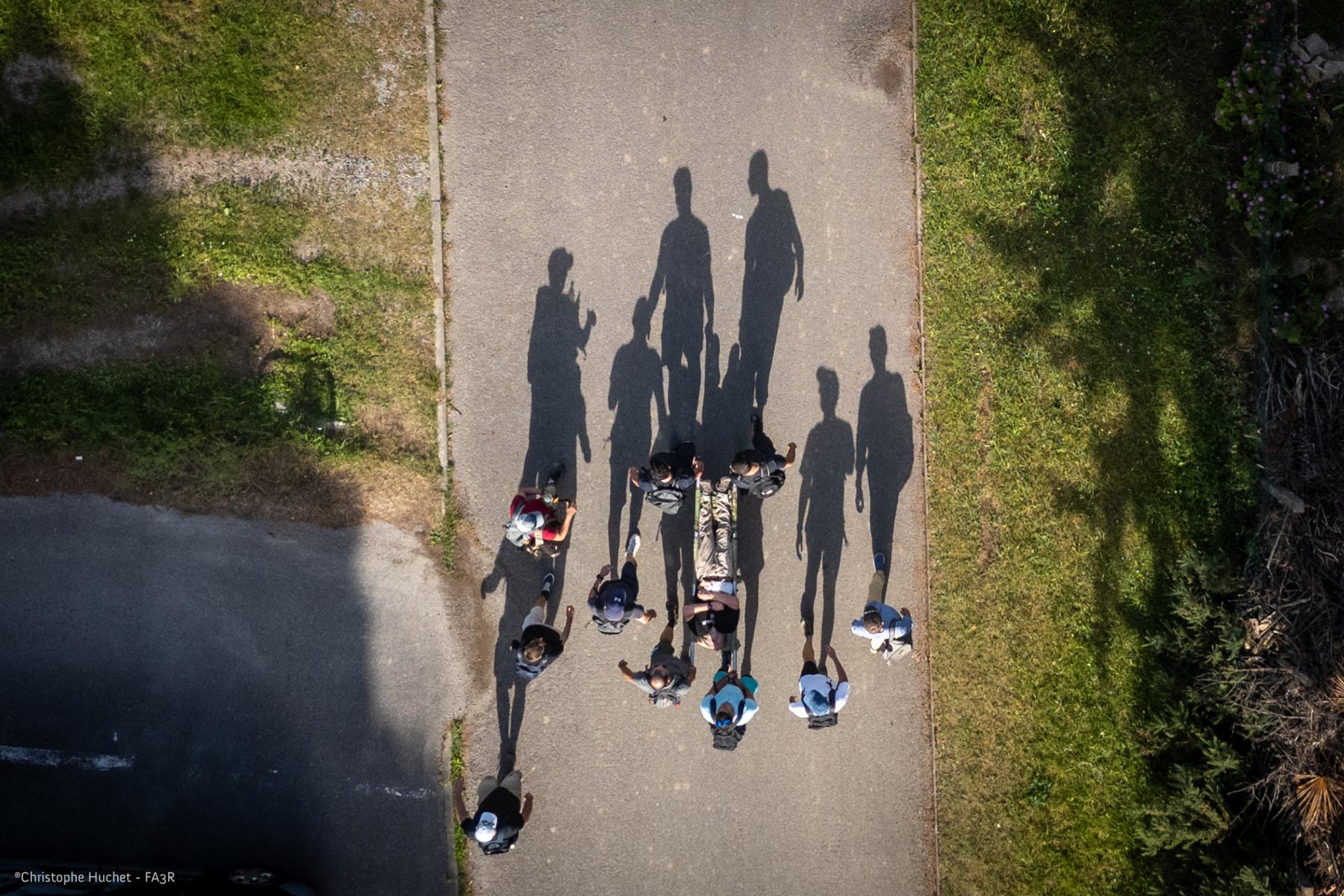 Vue drone d’une équipe portant un brancard. Ombres allongées sur le bitume et bordures d’herbe sous un soleil intense.