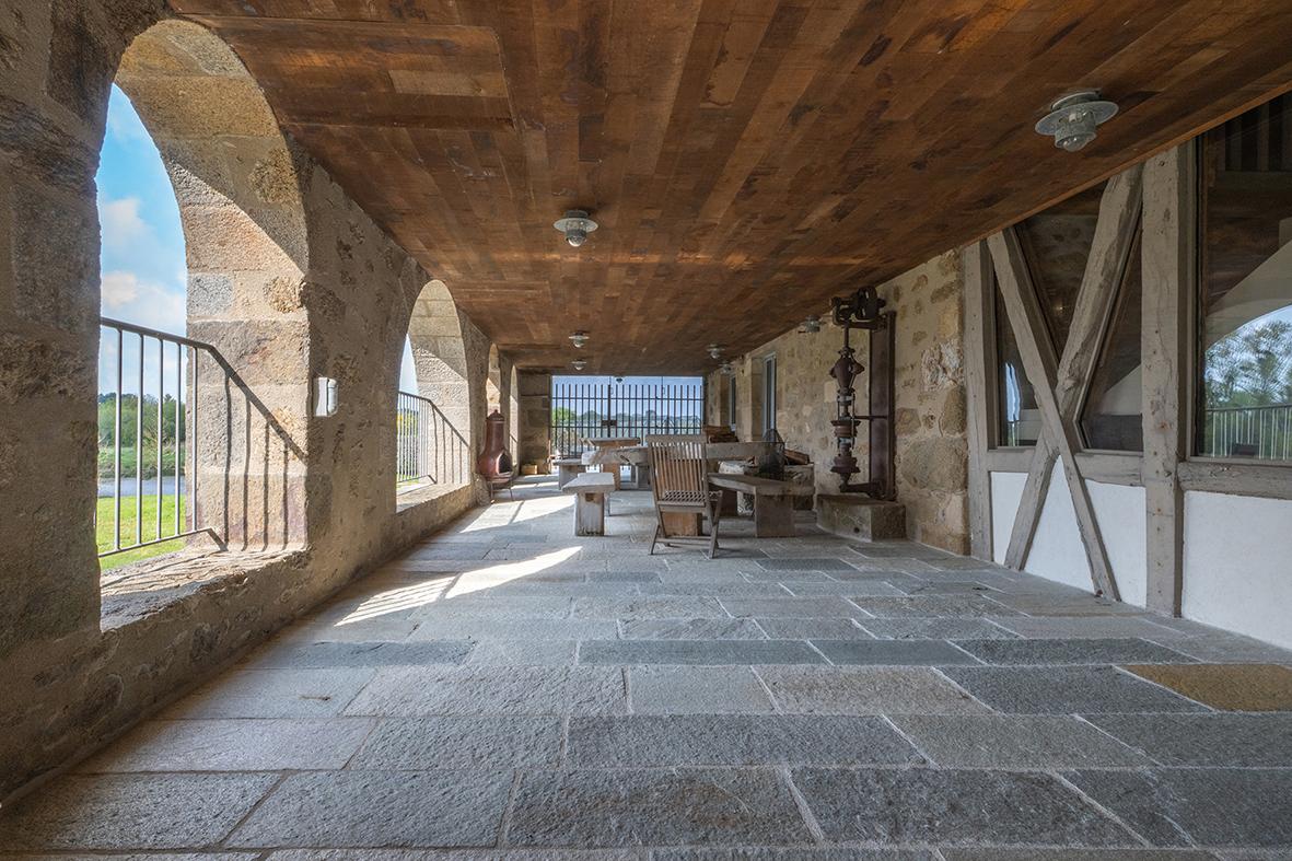 Terrasse abritée sous des arcades en pierre avec plafond en bois massif, sol en dalles de granit et mobilier rustique.