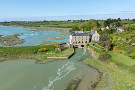 Moulin à marée bordant un estuaire verdoyant sous un ciel bleu clair en vue aérienne par drone.