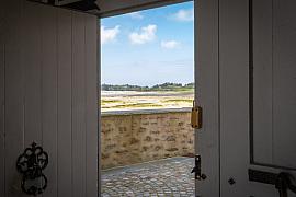 Porte blanche ouverte d’un moulin à marée révélant une cour pavée et un paysage littoral breton sous ciel bleu.