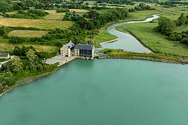 Vue aérienne du Moulin de Rochegoude entouré de verdure au bord d’une eau turquoise sous un ciel clair de Bretagne.